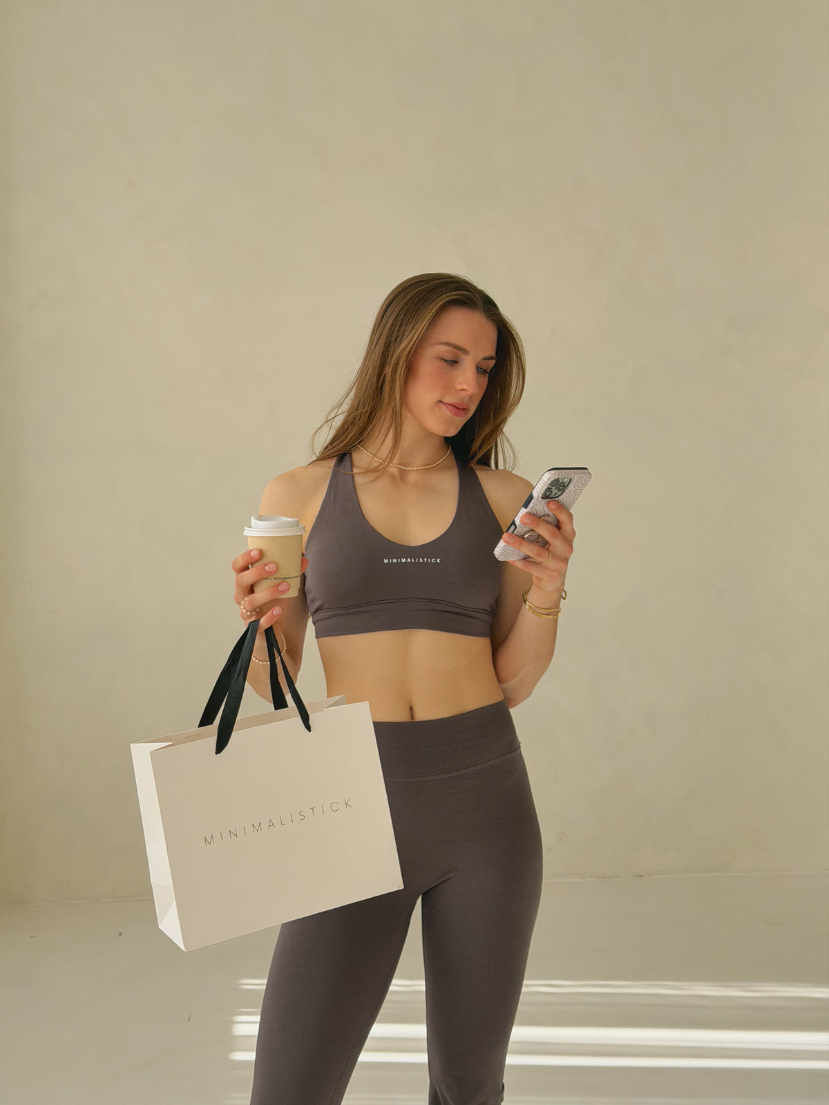 Modelka v bavlněné sportovní podprsence a legínách drží kávu, telefon a nákupní tašku MINIMALISTICK.